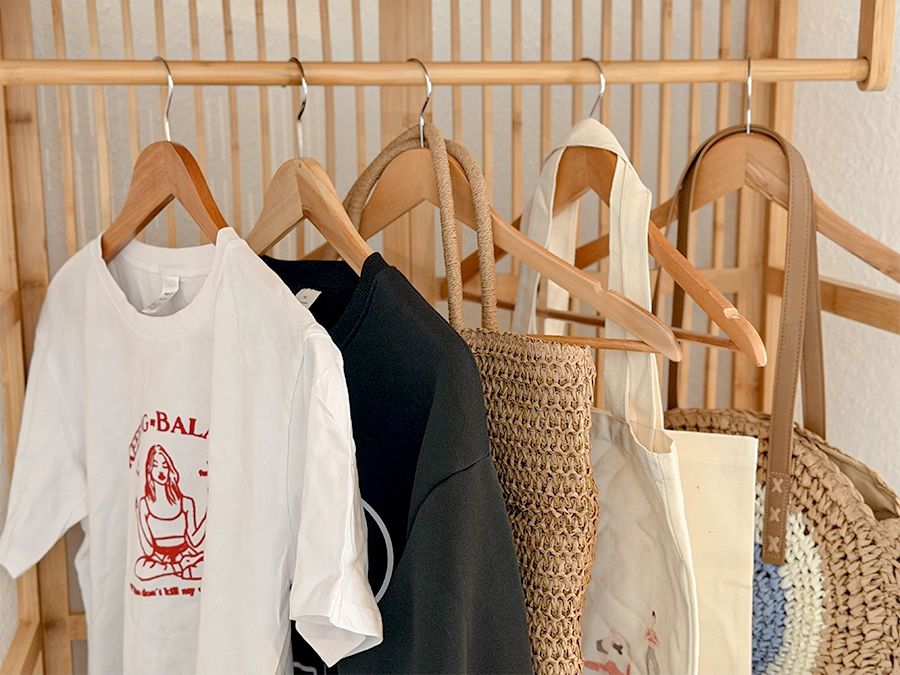 Auf einem hölzernen Kleiderständer in einem Mietatelier hängen ein weißes grafisches T-Shirt, ein schwarzes Oberteil, eine geflochtene Tasche, eine beigefarbene Tragetasche und eine runde Strohtasche ordentlich auf hölzernen Bügeln.