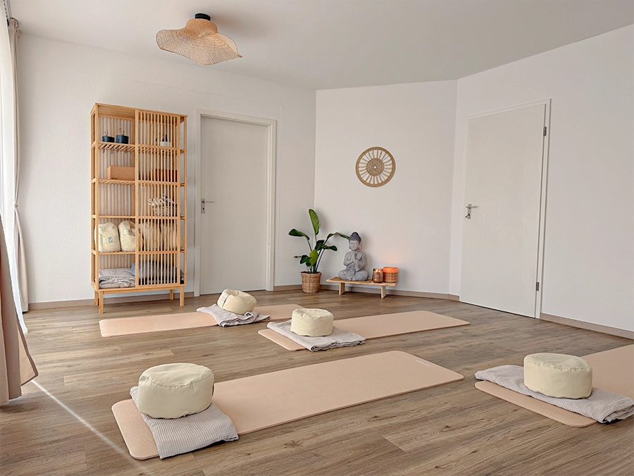 Ein ruhiges Yogastudio mit vier Matten, die jeweils mit einem runden Kissen und einer gefalteten Decke ausgelegt sind. Auf einem Holzregal stehen Handtücher, eine Topfpflanze und eine kleine Buddha-Statue in einem hellen, minimalistischen Raum mit Holzfußboden.
