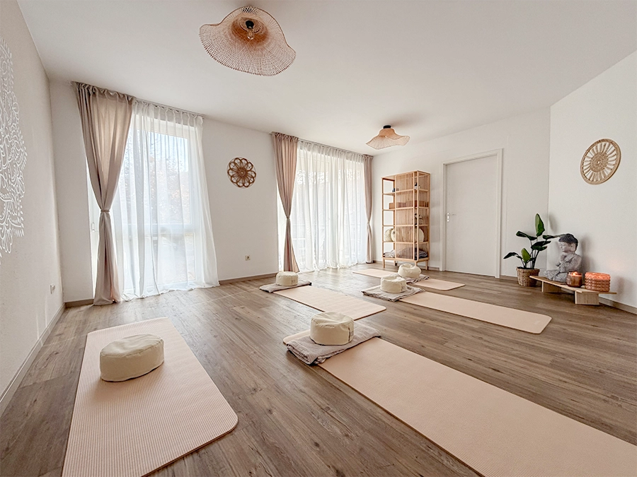 Ein helles, ruhiges Yogastudio mit Holzböden, großen Fenstern, die mit transparenten Vorhängen, sanfter Beleuchtung und ordentlich angeordneten Yogamatten drapiert sind. Studio mieten für Ihre nächste Sitzung und genießen Sie die beruhigende Atmosphäre mit Kerzen, Pflanzen und gemütlichen Kissen.