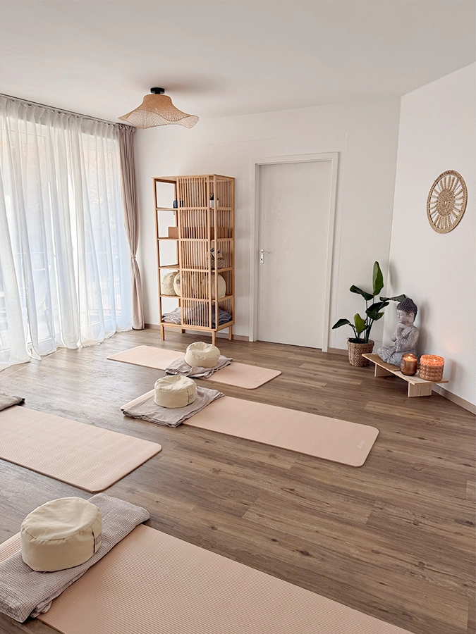 Ein helles, friedliches Yogastudio mit vier rosafarbenen Matten und Polstern auf einem Holzboden zum Mieten. Ein Bambusregal, Topfpflanzen, eine Buddha-Statue, Kerzen und transparente Vorhänge füllen den Raum mit Ruhe und natürlichem Licht.