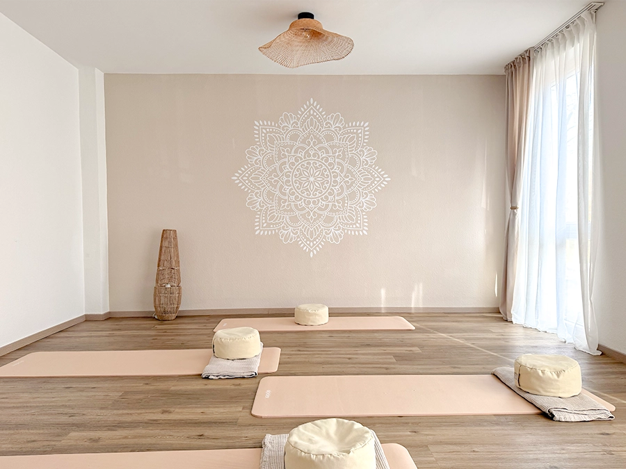 Ein helles, minimalistisches Yogastudio mit hellem Holzboden, cremefarbenen Yogamatten und runden Kissen, die auf dem Boden liegen. Ein großes Mandala schmückt die Rückwand - perfekt, wenn Sie ein Studio mieten möchten, um in Ruhe zu üben und natürliches Licht zu genießen.