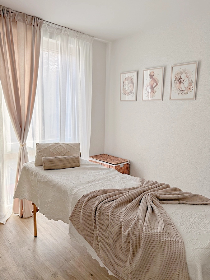 Ein heller, luftiger Massageraum mit sanftem Tageslicht, einem Massagetisch mit weißem Tuch und beiger Decke, dekorativen Kissen, leichten Vorhängen und drei gerahmten Kunstdrucken an einer weißen Wand.