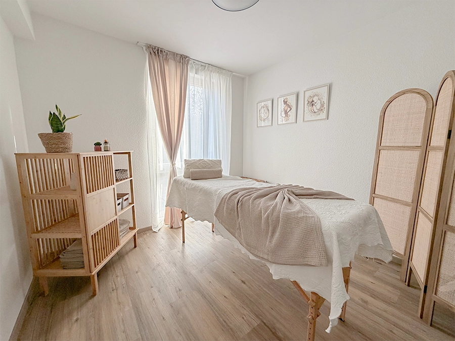 Ein heller, minimalistischer Massageraum, der dem Studio mieten zur Verfügung steht, mit einem hölzernen Massagetisch mit beiger Bettwäsche, einem Regal mit Handtüchern und Pflanzen, hellen Vorhängen, gerahmter Kunst an der Wand und einem klappbaren Raumteiler auf einem Holzboden.