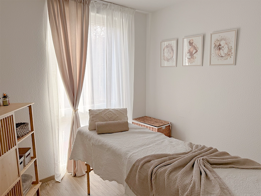 Ein gemütlicher, sonnendurchfluteter Raum, der sich perfekt zum Studiomieten eignet, mit einem sauber gefertigten Massagetisch, neutralen Decken, hellen Vorhängen, Wandkunst über einem Korbmöbel und einem kleinen Regal mit Dekoration in der Ecke.