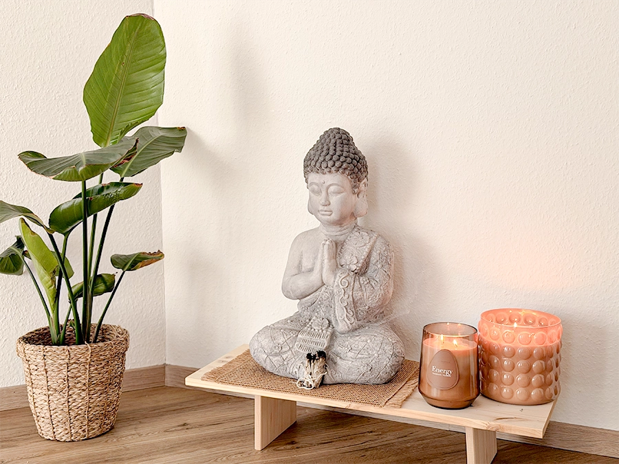 Eine kleine Buddha-Statue steht auf einem Holzpodest neben einer brennenden braunen Kerze und einem dekorativen Kerzenhalter. Eine grüne Topfpflanze in einem geflochtenen Korb ist perfekt für diejenigen, die ein Studio mieten möchten, und bringt Ruhe in die Einrichtung auf dem Holzboden.