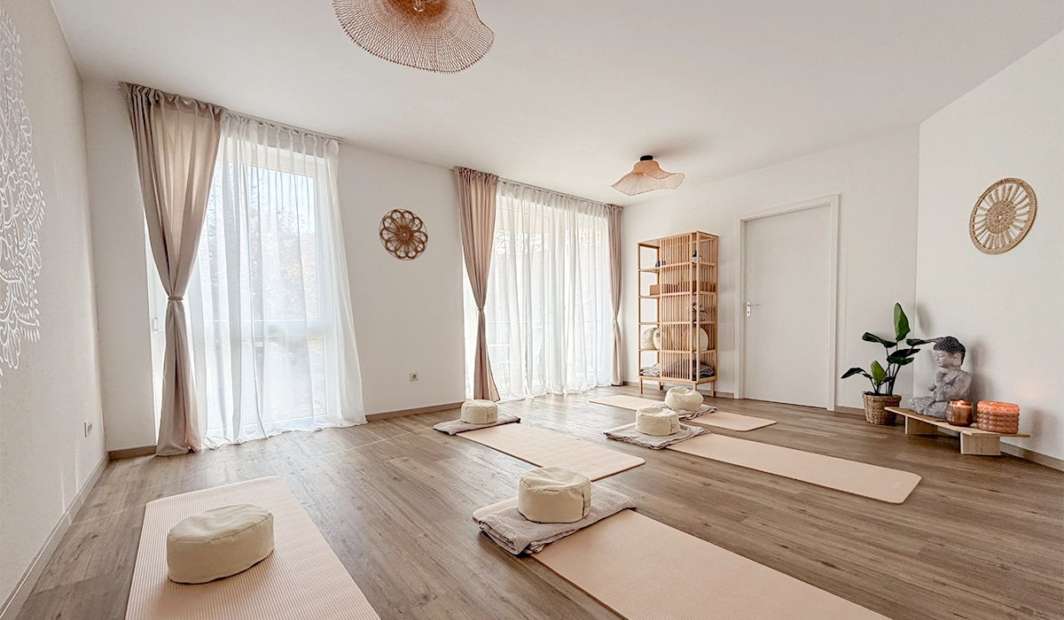 Ein heller freundlicher raum mit weissen und beigen wänden. Auf dem Boden liegen Yoga Matten und Kissen. Ein Buddha ist in der Ecke auf einem Holzbrett.