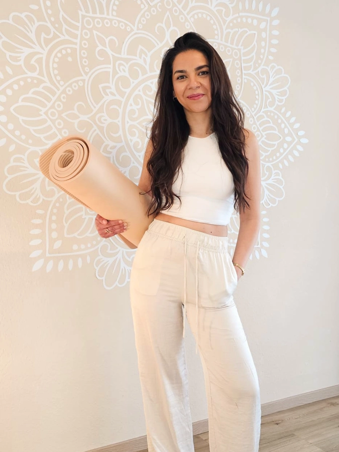 Kursleiterin für schwangerenyoga steht vor einer wand mit einem Tribal und hält eine Yoga Matte unterm Arm